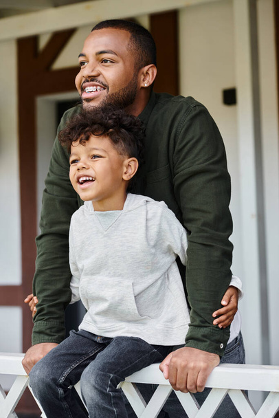 gioioso ragazzo afroamericano seduto sul portico e abbracciare il padre in bretelle sul cortile di casa - Foto, immagini