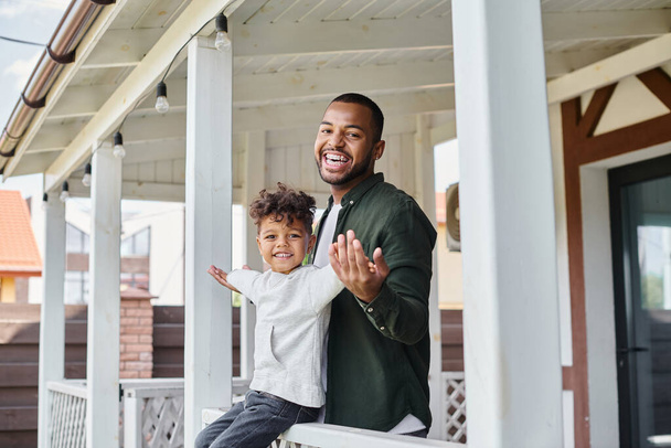 momentos felices, niño afroamericano alegre sentado en el porche y cogido de la mano con el padre en frenos - Foto, imagen