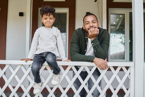 joyful african american father and son smiling and sitting on porch of house, family portrait - Foto, Bild