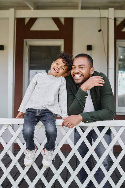 positive african american father and son smiling and sitting on porch of house, family portrait - Фото, изображение