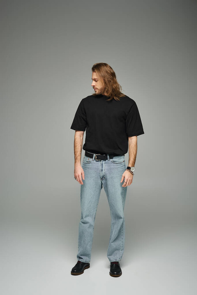 longitud completa del hombre guapo barbudo en camiseta negra y pantalones vaqueros de pie sobre fondo gris - Foto, Imagen