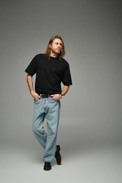 full length of bearded handsome man in black t-shirt and jeans standing with hand in pockets on grey - Photo, Image