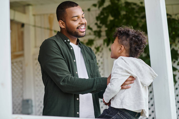 Neşeli Afro-Amerikan babası banliyödeki verandada oturan tatlı kıvırcık oğluna sarılıyor. - Fotoğraf, Görsel