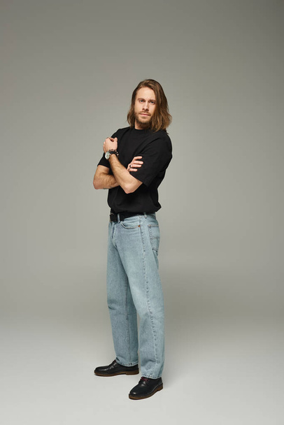 full length of bearded and handsome man with long hair posing in jeans and t-shirt on grey backdrop - Foto, afbeelding