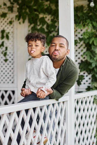heureux père afro-américain et fils bouclé collant la langue tout en étant assis près de la clôture blanche - Photo, image