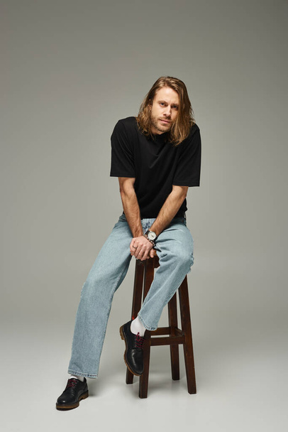 full length of bearded and handsome man with long hair sitting in jeans and t-shirt on high stool - Fotoğraf, Görsel