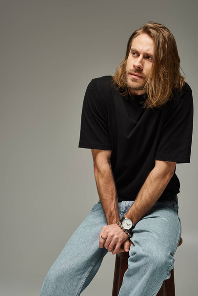 portrét vousatého a pohledného muže s dlouhými vlasy v džínách a tričku sedícího na židli na šedé - Fotografie, Obrázek