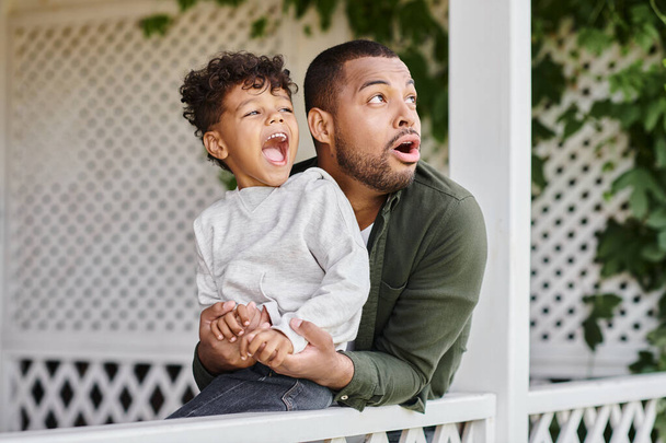 astonished african american father and son looking away surprisingly while sitting near white fence - Фото, изображение