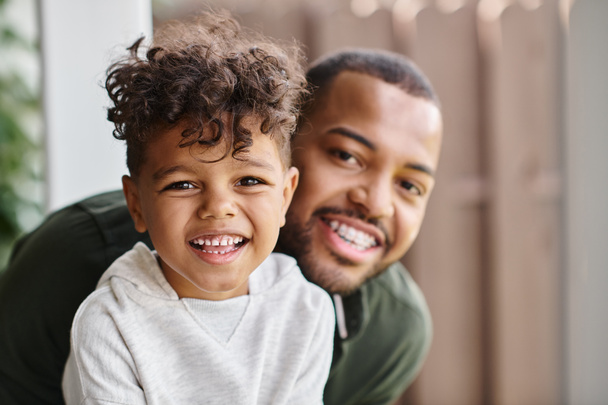 Familienporträt eines fröhlichen afrikanisch-amerikanischen Mannes mit Zahnspange, der seinen lockigen Sohn umarmt, während er in die Kamera schaut - Foto, Bild