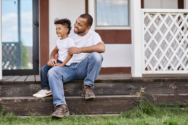 homem americano africano alegre em aparelho abraçando filho encaracolado enquanto sentado no alpendre da casa moderna - Foto, Imagem