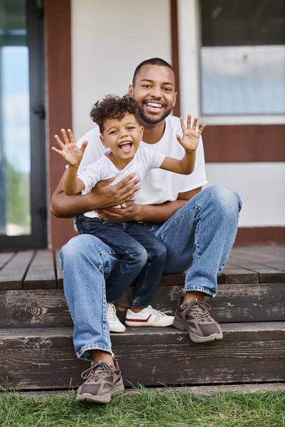 vidám afro-amerikai férfi fogszabályzóval öleli izgatott fia, miközben ül a tornácon a modern ház - Fotó, kép