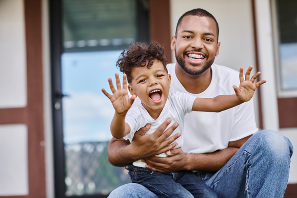 幸せなアフリカ系アメリカ人男性は,現代の家のポーチに座っている間興奮する息子を抱きしめます - 写真・画像