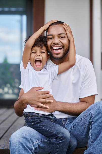excited african american boy hugging happy father in braces while sitting on porch of modern house - Photo, Image