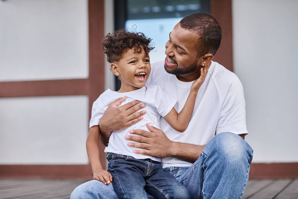 alegre afroamericano chico abrazando feliz padre en aparatos ortopédicos mientras sentado en porche de casa moderna - Foto, Imagen