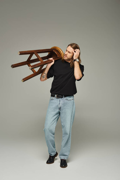 Vollbärtiger Mann mit langen Haaren posiert in Jeans und T-Shirt, während er einen hohen Stuhl trägt - Foto, Bild
