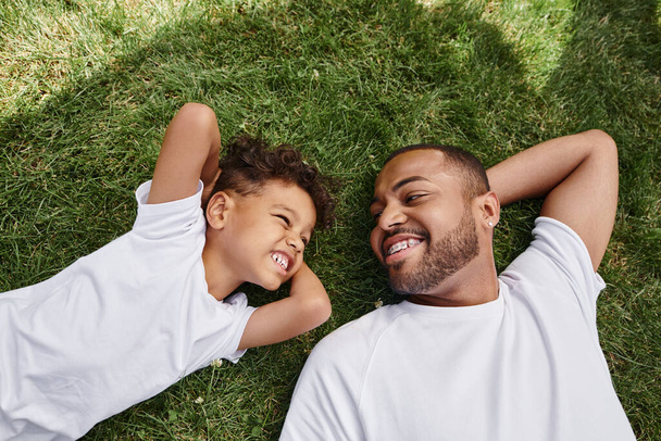 top view of cheerful african american father and cute son lying on green lawn, family photo - Photo, Image