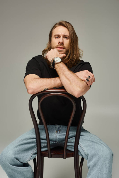 bearded and good looking man with long hair sitting in jeans and t-shirt on wooden chair on grey - Fotoğraf, Görsel