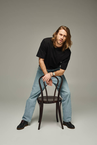 full length of bearded good looking man with long hair in jeans and t-shirt sitting on chair on grey - Fotoğraf, Görsel