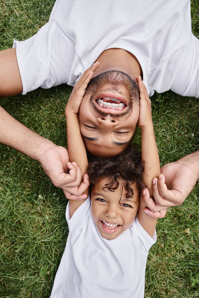 アフリカ系アメリカ人の父親と息子が裏庭に緑の芝生に横たわり,幸せな家族の思い出を思い出します. - 写真・画像