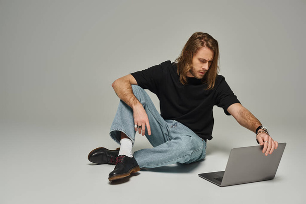 bell'uomo con i capelli lunghi e la barba seduto in jeans denim sul pavimento e utilizzando il computer portatile su grigio - Foto, immagini