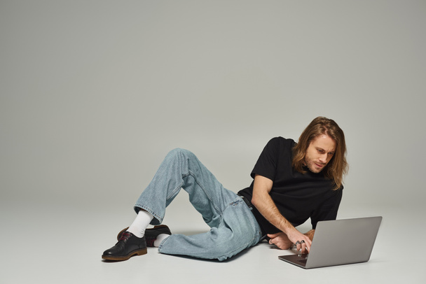 schöner Mann mit langen Haaren und Bart sitzt in Jeans auf dem Boden und benutzt Laptop auf grau - Foto, Bild