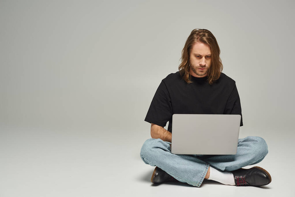 красивий чоловік з довгим волоссям і бородою сидить з схрещеними ногами на підлозі і використовує ноутбук на сірому - Фото, зображення