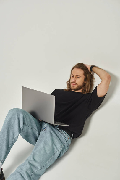 Uzun saçlı ve sakallı yakışıklı adam beyaz duvara yaslanıyor ve gri arka planda dizüstü bilgisayar kullanıyor. - Fotoğraf, Görsel