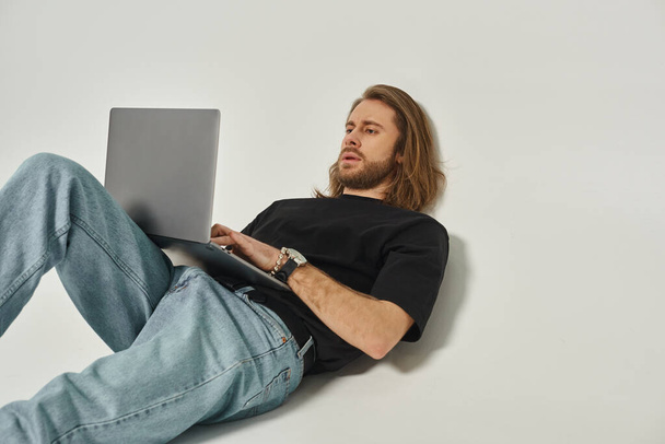 Uzun saçlı ve sakallı yakışıklı adam beyaz duvara yaslanıyor ve gri arka planda dizüstü bilgisayar kullanıyor. - Fotoğraf, Görsel
