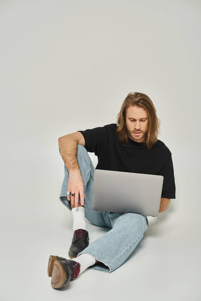 gutaussehende Freiberuflerin mit langen Haaren und Bart sitzt auf dem Boden und benutzt Laptop auf grauem Hintergrund - Foto, Bild