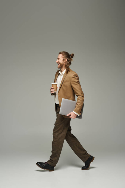 happy businessman with beard and long hair holding coffee to go and laptop while walking on grey - Foto, imagen