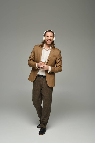 グレーの背景にコーヒーを持っているヘッドフォンの陽気なひげ付きビジネスマン,正式な服装 - 写真・画像
