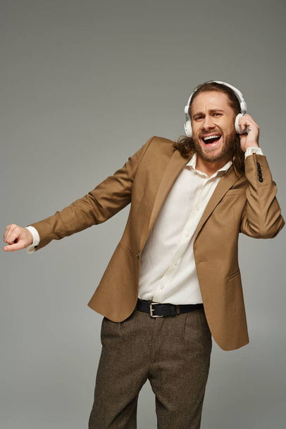 izgatott és szakállas üzletember fülhallgatóban zenét hallgat szürke háttérrel, hivatalos öltözékben - Fotó, kép
