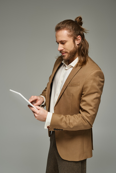 brodaty biznesmen z długimi włosami pozujący w formalnym stroju i korzystający z tabletu cyfrowego na szarym tle - Zdjęcie, obraz