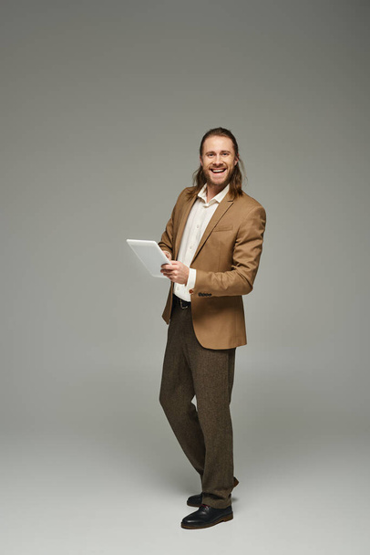 glücklicher Geschäftsmann mit langen Haaren posiert in formeller Kleidung und mit digitalem Tablet vor grauem Hintergrund - Foto, Bild