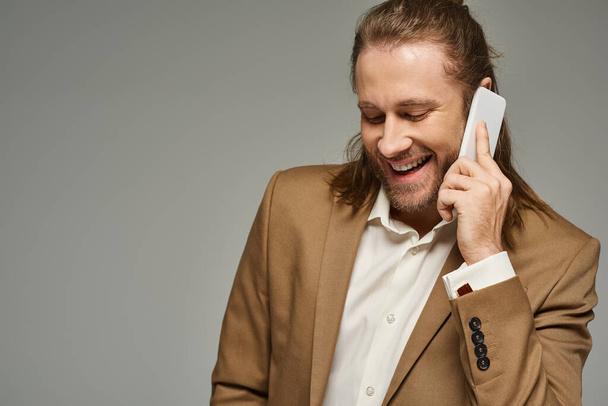 gutaussehender und glücklicher bärtiger Geschäftsmann in formeller Kleidung, der vor grauem Hintergrund mit dem Smartphone spricht - Foto, Bild