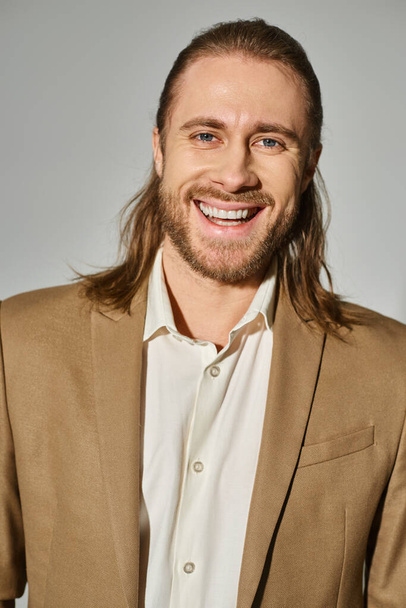 portrait of positive businessman with long hair and beard posing in beige blazer on grey backdrop - Foto, imagen