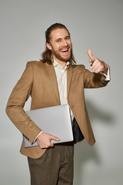 vrolijk zakenman in beige formele kleding houden laptop en tonen duim omhoog op grijze achtergrond - Foto, afbeelding