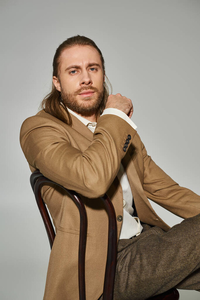 good looking bearded businessman in beige formal attire sitting on wooden chair on grey background - Fotografie, Obrázek