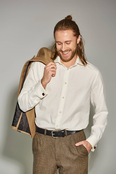 позитивный и бородатый бизнесмен в белой рубашке позирует с бежевым блейзером на плече на сером - Фото, изображение