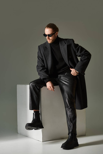 full length, fashionable model in trendy sunglasses and coat sitting on white cube on grey backdrop - Foto, imagen