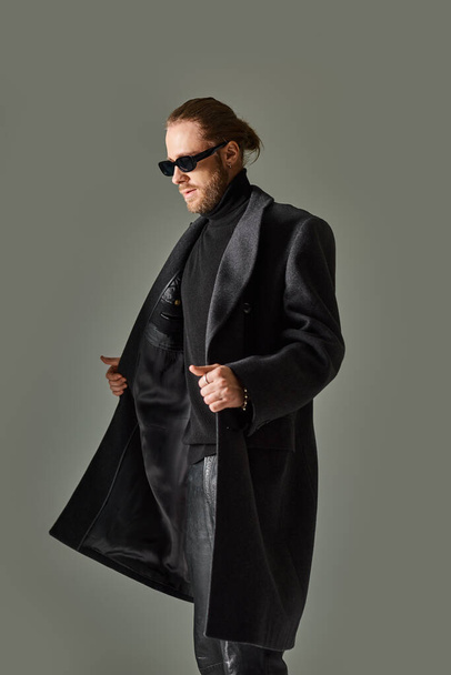 portrait of handsome bearded man in trendy sunglasses and black attire walking on grey background - Foto, imagen