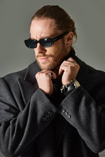 good looking bearded man in trendy sunglasses and black attire adjusting collar of turtleneck - Photo, Image
