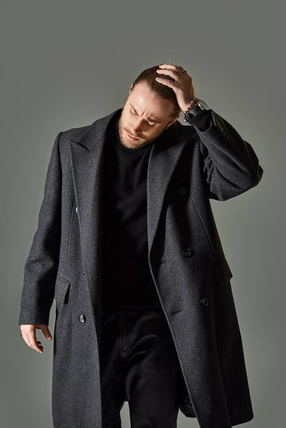 jóképű és szakállas férfi divatos fekete öltözet és gyapjú kabát pózol szürke háttér - Fotó, kép