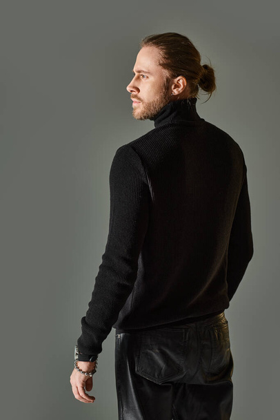 bearded handsome man in black turtleneck sweater and leather pants standing on grey background - Photo, Image