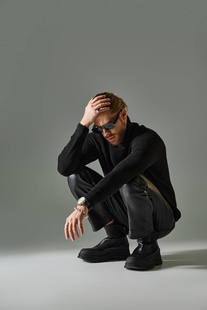 bearded man in stylish sunglasses, turtleneck sweater and leather pants sitting on haunches on grey - Photo, Image