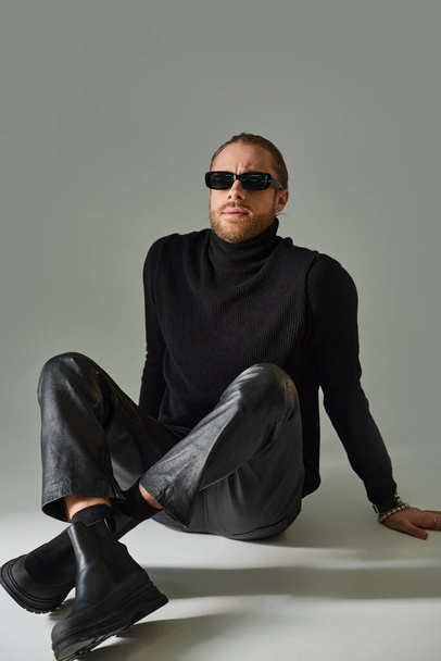 handsome man in sunglasses, turtleneck sweater and leather pants sitting with crossed legs on grey - Foto, Imagem