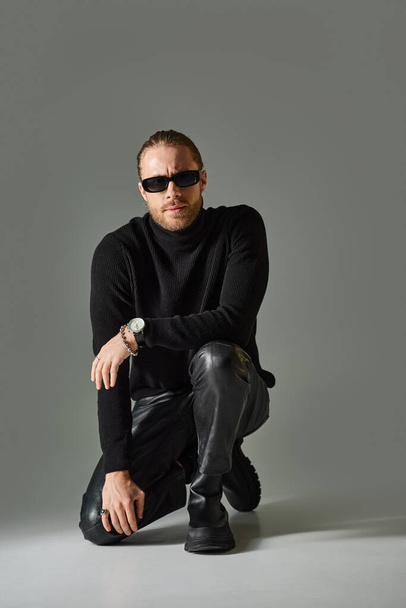 bearded man in trendy sunglasses, turtleneck sweater and leather pants sitting on grey background - Фото, зображення