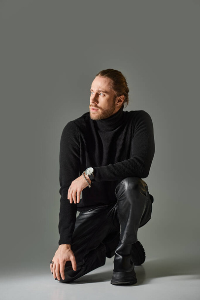 full length of bearded man in turtleneck sweater and black leather pants sitting on grey background - Fotografie, Obrázek