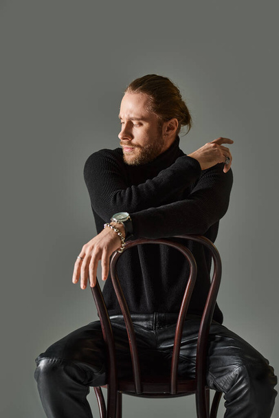 bearded man in turtleneck and black leather pants sitting on wooden chair on grey background - Fotoğraf, Görsel