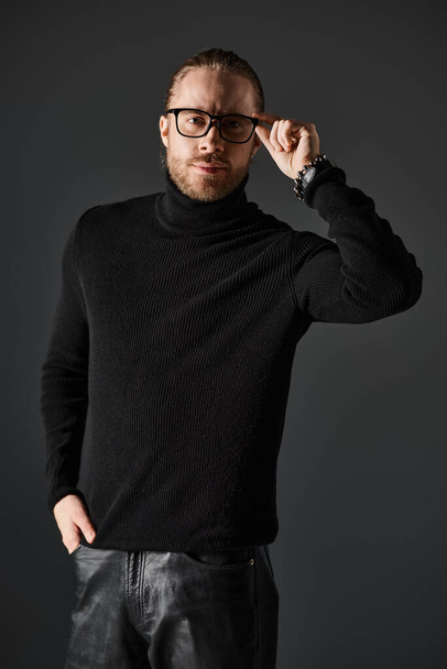 handsome man in turtleneck sweater and eyeglasses standing in leather pants on grey background - Foto, afbeelding